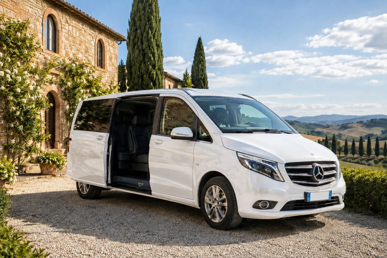 A white nine-seat Mercedes van on the gravel drive of a Tuscan villa, side door open, cypresses and hills behind