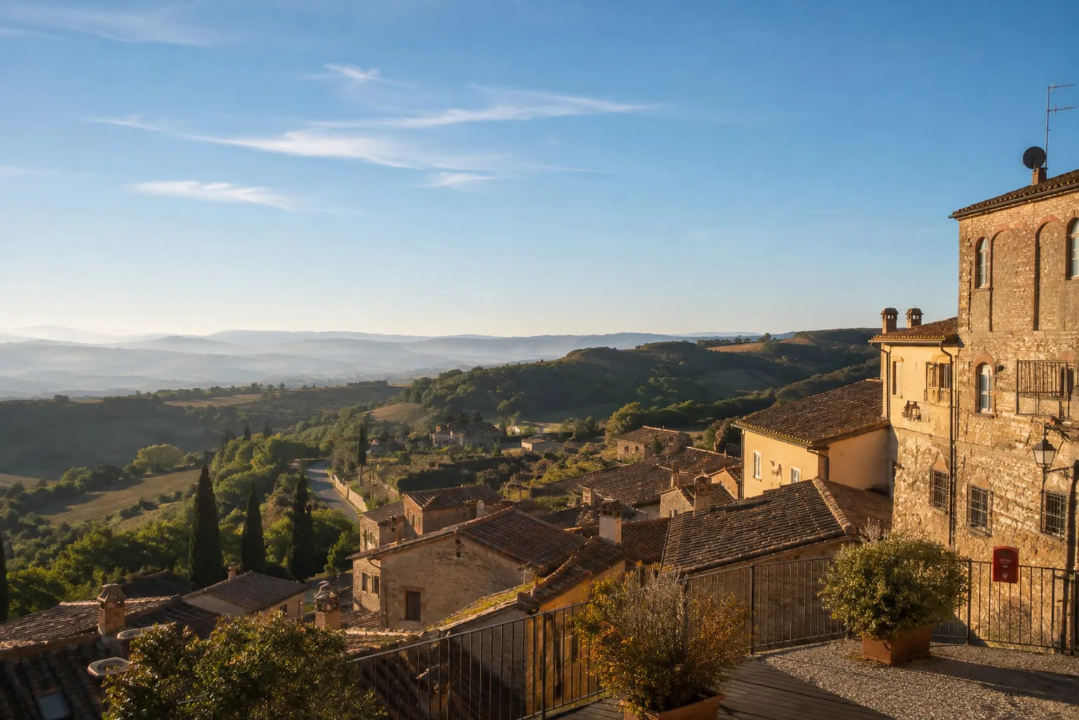 A terrace above the terracotta rooftops of a Tuscan hill village, looking out over wooded slopes to hills fading into haze in low warm light